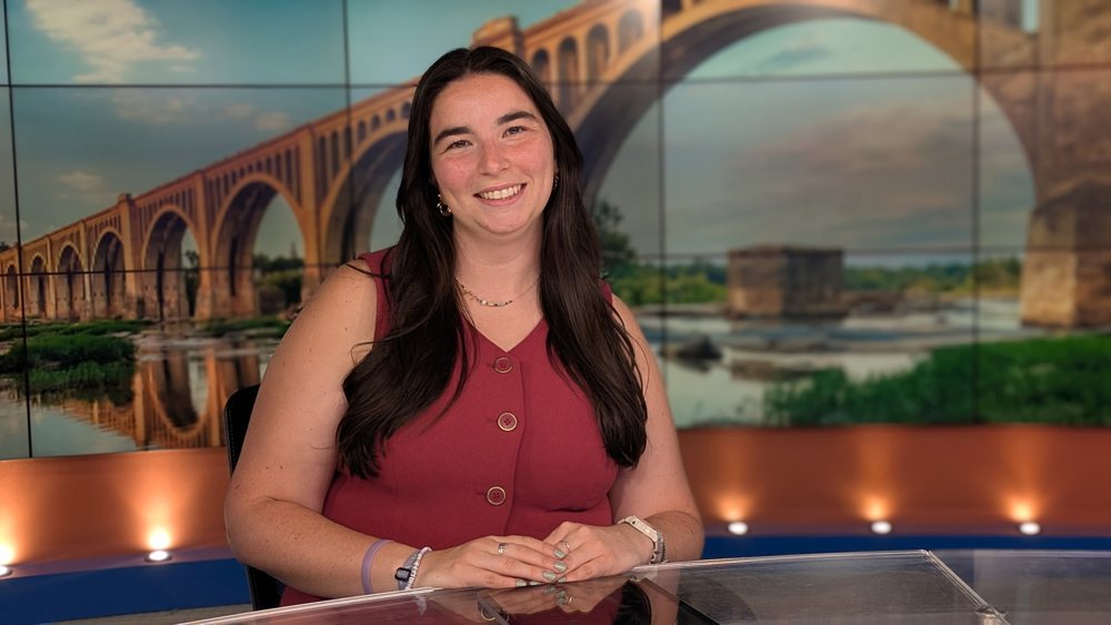 Headshot of Amory Doyle smiling at a news desk in front of a bridge backdrop