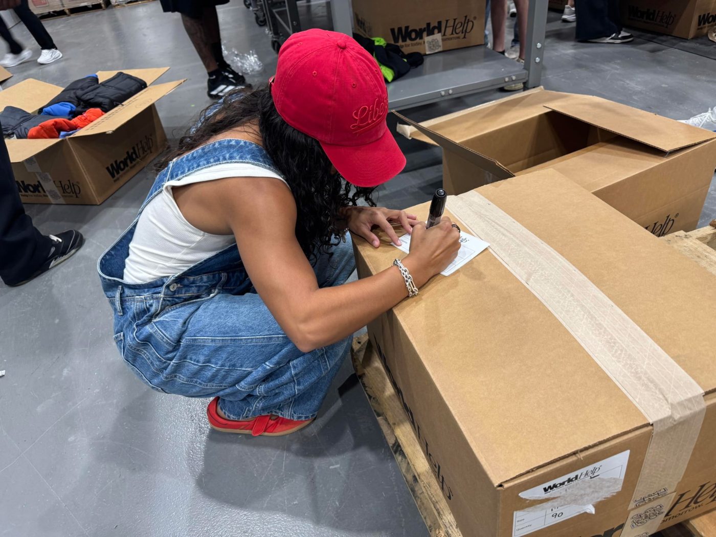 A volunteer in a red cap crouching to write on a label on a taped cardboard box